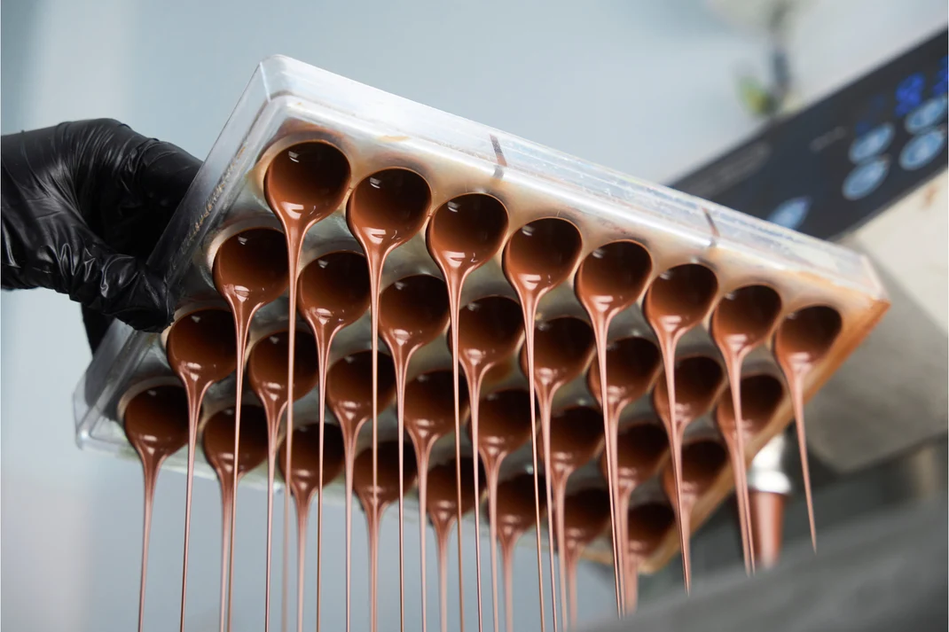 A gloved hand lifting a polycarbonate bonbon mould, tempered chocolate draining from every cavity in long threads