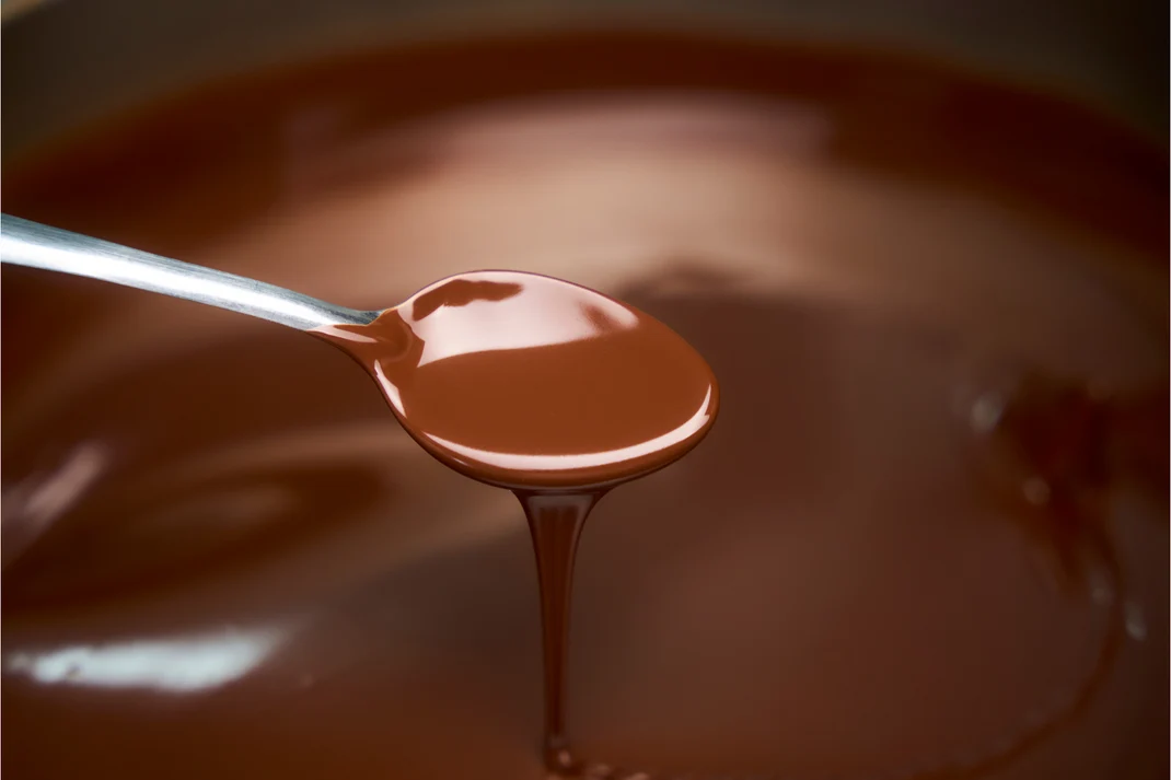 A spoon lifting glossy melted chocolate from the tempering bowl